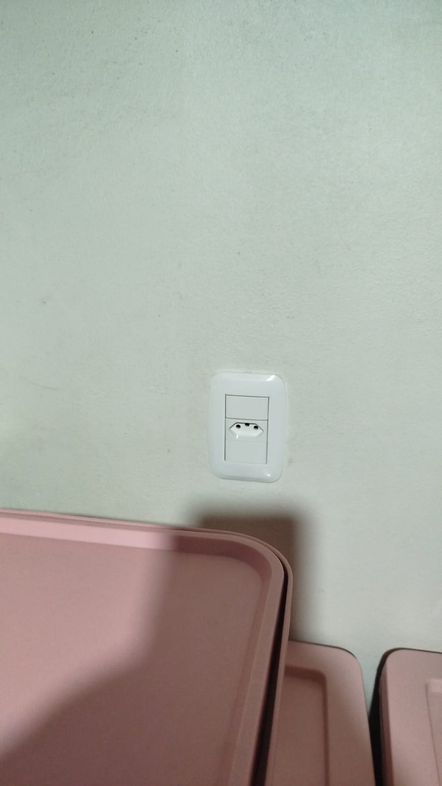 tomada elétrica branca com interruptor numa parede clara, com peças rosa plásticas em primeiro plano embaixo 